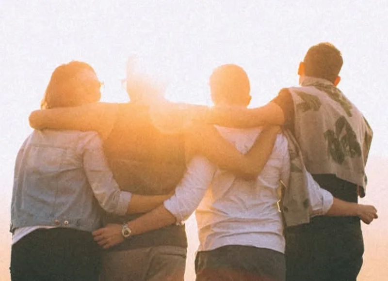 Four friends with arms around each other watching the sun set over hills